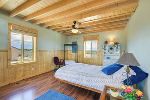 Ein Schlafzimmer mit Bett, Deckenventilator, Fenster, Stuhl und Nachttisch mit Lampe und Blumen.