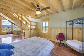 A bedroom with a bed, a chair, a bunk bed, wooden walls, a ceiling fan, and large windows. - Sangre de Cristo Views! Retreat Near Wild Horses! (San Luis)