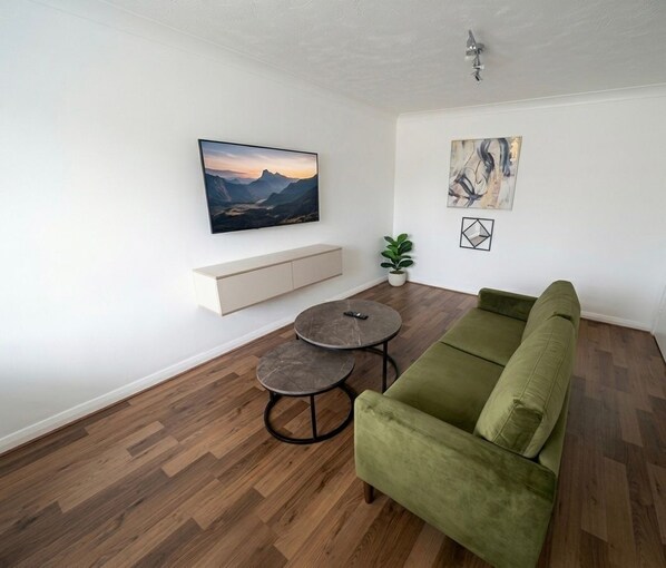 A living room with a green sofa, a round coffee table, a mounted TV, a plant, and artwork on the walls. - Keepers Court Luxury Stay (South Croydon)