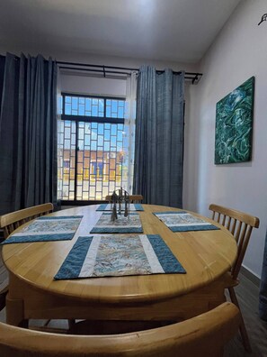 A dining area with a round wooden table, four chairs, a window with curtains, and a painting on the wall. - Azure Chameleon House (Zanzibar)