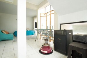 Um quarto com chão de azulejos, um sofá azul, uma mesa de vidro com cadeiras, um refrigerador preto e um aquecedor.