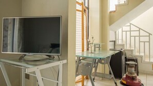 A room with a television on a glass table, a window with blinds, and a staircase with a geometric design.