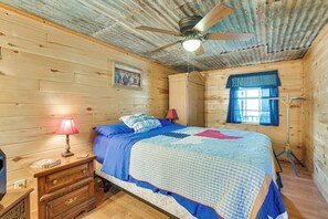 Un dormitorio de cabaña de madera con cama, mesita de noche, lámpara, ventilador de techo y ventana con cortinas.