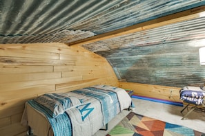 A room with a wooden bench, a patterned blanket, a colorful rug, and a corrugated metal ceiling.