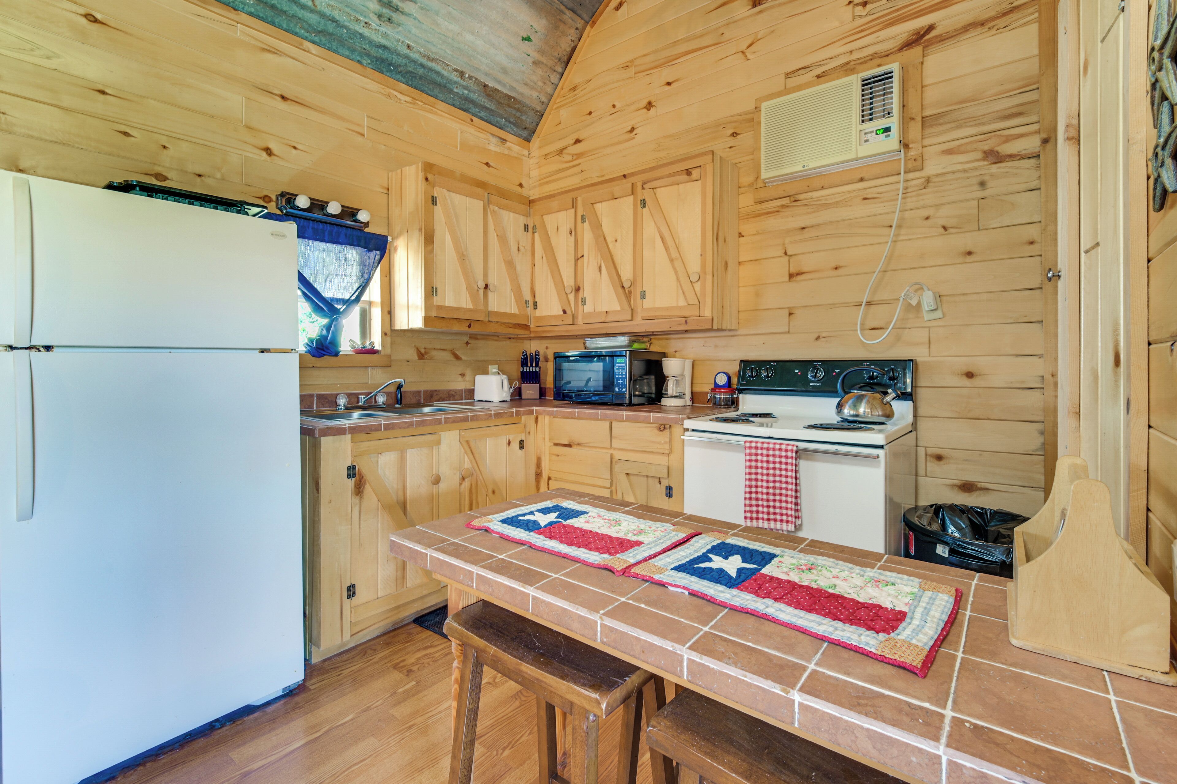 Una cocina con armarios de madera, un refrigerador, un microondas, una estufa y una mesa con colcha.