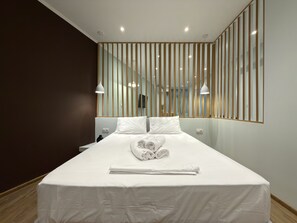Une chambre d’hôtel moderne avec un grand lit, du linge de lit blanc, un plancher en bois et une vue sur l’intérieur à travers une cloison à rayures.