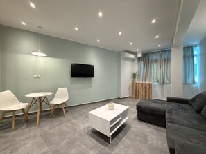A modern living room with a flat-screen TV, a coffee table, two white chairs, a dark sofa, and a wooden side table with a plant. - SmartLiving Central Apt 11 (Thessaloniki)