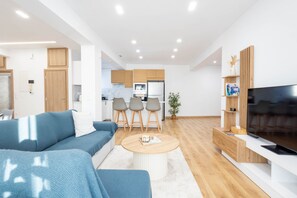 Uma sala de estar moderna com sofá, mesa de centro, TV e área de cozinha ao fundo.