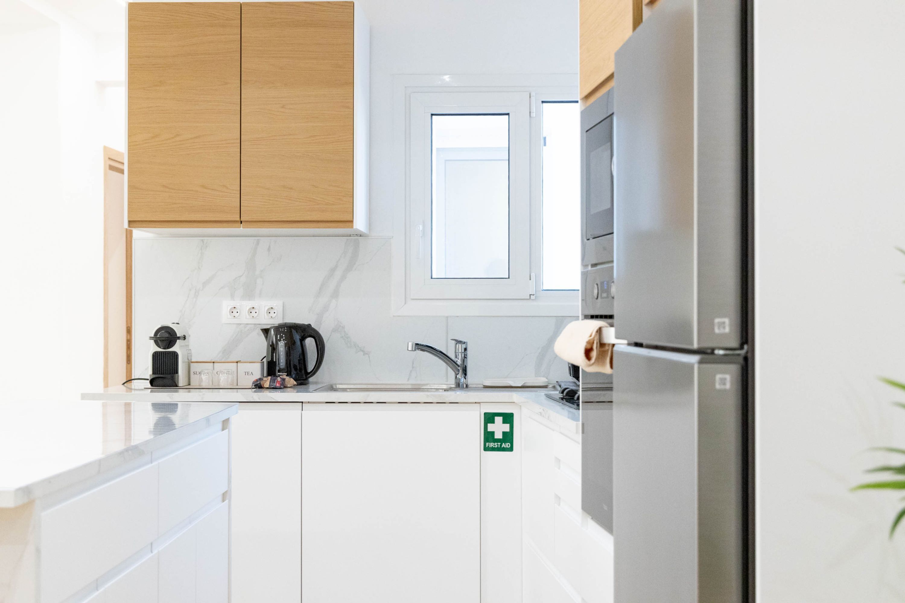 A modern kitchen with white cabinets, a stainless steel refrigerator, a coffee maker, and a first aid sign.