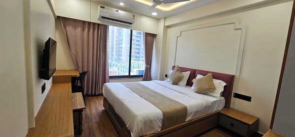 A hotel room with a bed, bedside table, TV, air conditioning unit, window with curtains, and a view of buildings. - Hotel Silvera Inn (Ahmedabad)