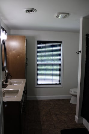 A bathroom with a toilet, sink, window with blinds, and a shower area.