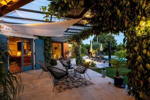 A covered outdoor seating area with chairs and a table, a pool in the background, and a glass door entrance.