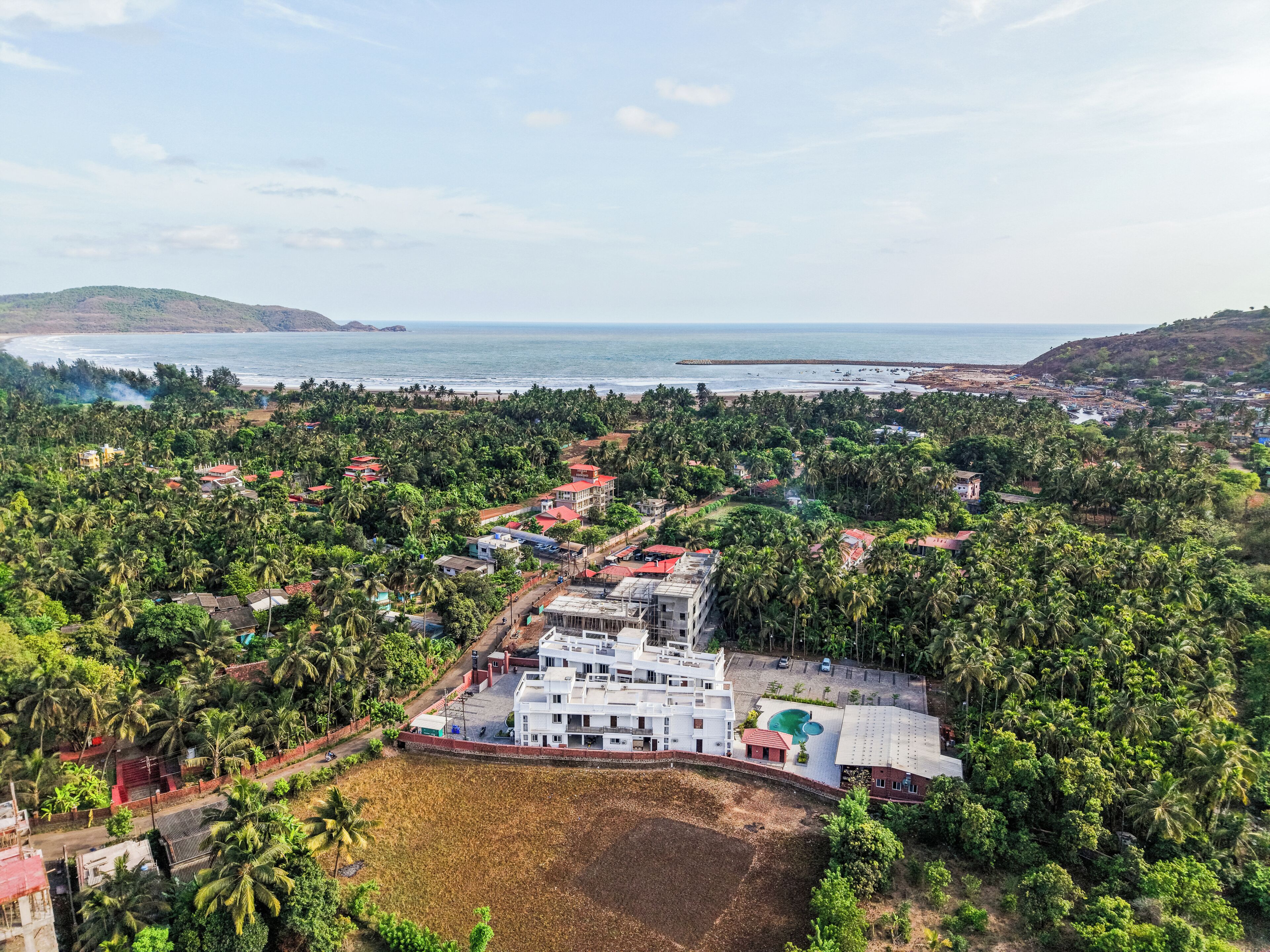 An aerial view of a coastal town with a prominent white building, dense greenery, and a beach in the background.