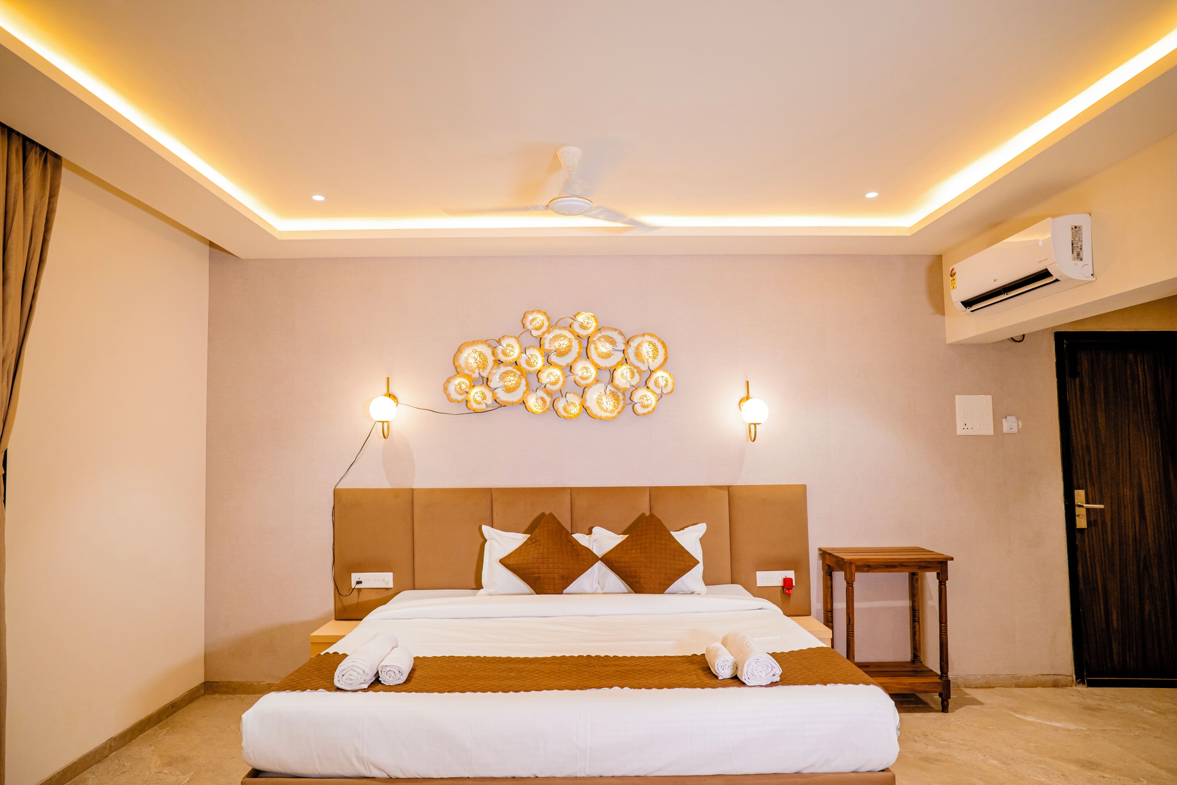 A hotel room with a bed, bedside tables, an air conditioning unit, a ceiling fan, and a wall-mounted light fixture.