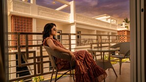 A woman seated on a wicker chair on a balcony with a view of a building with white brick walls and a balcony railing. - Breeze Inn Beach Resort (Shrivardhan)