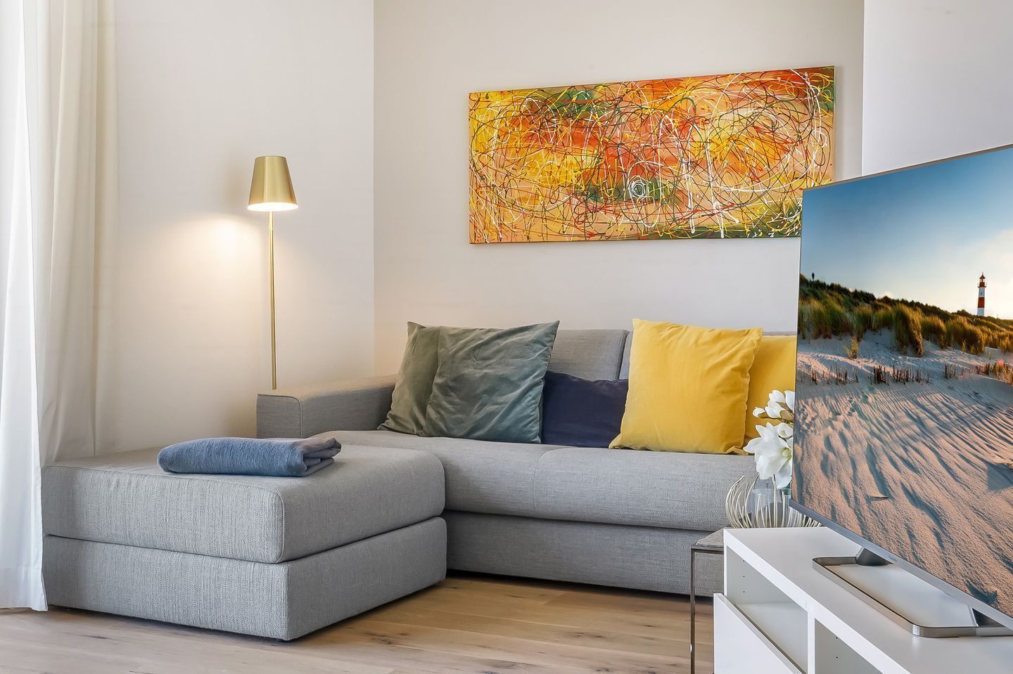 Een moderne woonkamer met een grijze modulaire bank, een vloerlamp, een kleurrijk abstract schilderij en een schilderij van een strandgezicht.