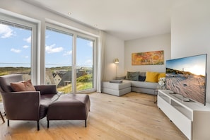 Een moderne woonkamer met een bank, een televisie en een groot raam met uitzicht op gebouwen en groen.
