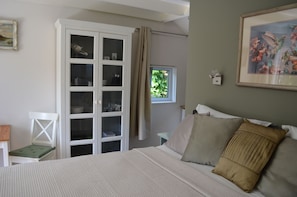 Ein Schlafzimmer mit einem Bett, einem weißen Kleiderschrank, einem Fenster mit Vorhängen, einem gerahmten Bild und einem kleinen Tisch mit einer Lampe.