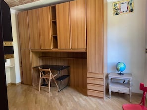 A room with a wooden wardrobe, a desk with a chair, a small table with a globe, and a red chair.