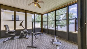 Ein überdachter Fitnessbereich im Freien mit Trainingsgeräten, großen Fenstern und einem Deckenventilator.