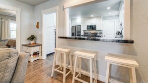 Cocina moderna con armarios blancos, un refrigerador de acero inoxidable y una encimera negra. Hay dos taburetes blancos y una pequeña mesa con una planta.
