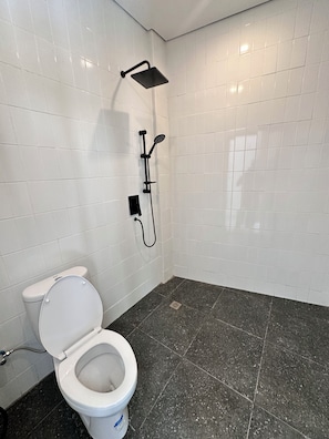 A bathroom with white tiled walls, a white toilet, a shower with a black hose, and a dark speckled floor. - Sedana Villa (Badung)