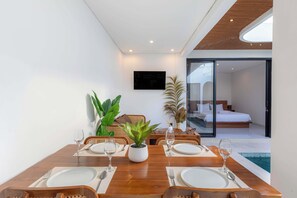 A dining area with a wooden table set for two, surrounded by potted plants, and a bedroom visible through a glass door. - Villa Didi by Sedana Cottage (Badung)