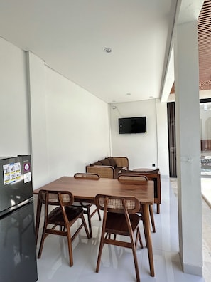 A modern dining area with a wooden table and chairs, a black refrigerator, and a flat-screen TV mounted on the wall. - Sedana Villa (Badung)