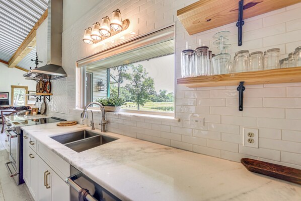 A kitchen with a large window, white tiled backsplash, stainless steel sink, and wooden shelves with jars.