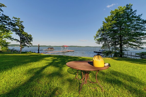 Une zone herbeuse avec une table en métal et un sac jaune, d’où l’on a vue sur un lac avec un quai et un petit bâtiment.