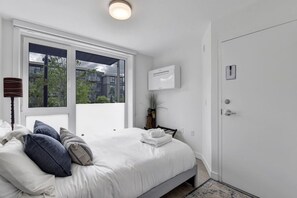 A bedroom with a bed, a window, a wall-mounted air conditioner, and a door. - Stunning One Bedroom Lonsdale Quay Eco-Suite (North Vancouver)