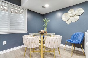 Un coin repas comprenant une table ronde en verre, quatre chaises rembourrées aux pieds dorés, une chaise bleue et un élément décoratif fixé au mur.