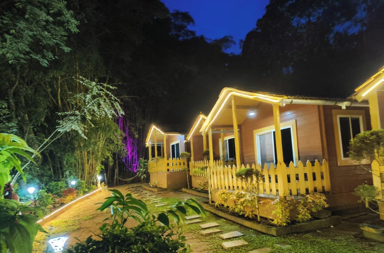 A row of wooden cabins with a picket fence, surrounded by lush greenery and illuminated by warm lights.
