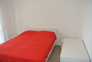 A single bed with a red bedspread in a room with white walls and a white nightstand. - Stylish Pool View Flat With two Terraces (Lignano Sabbiadoro)