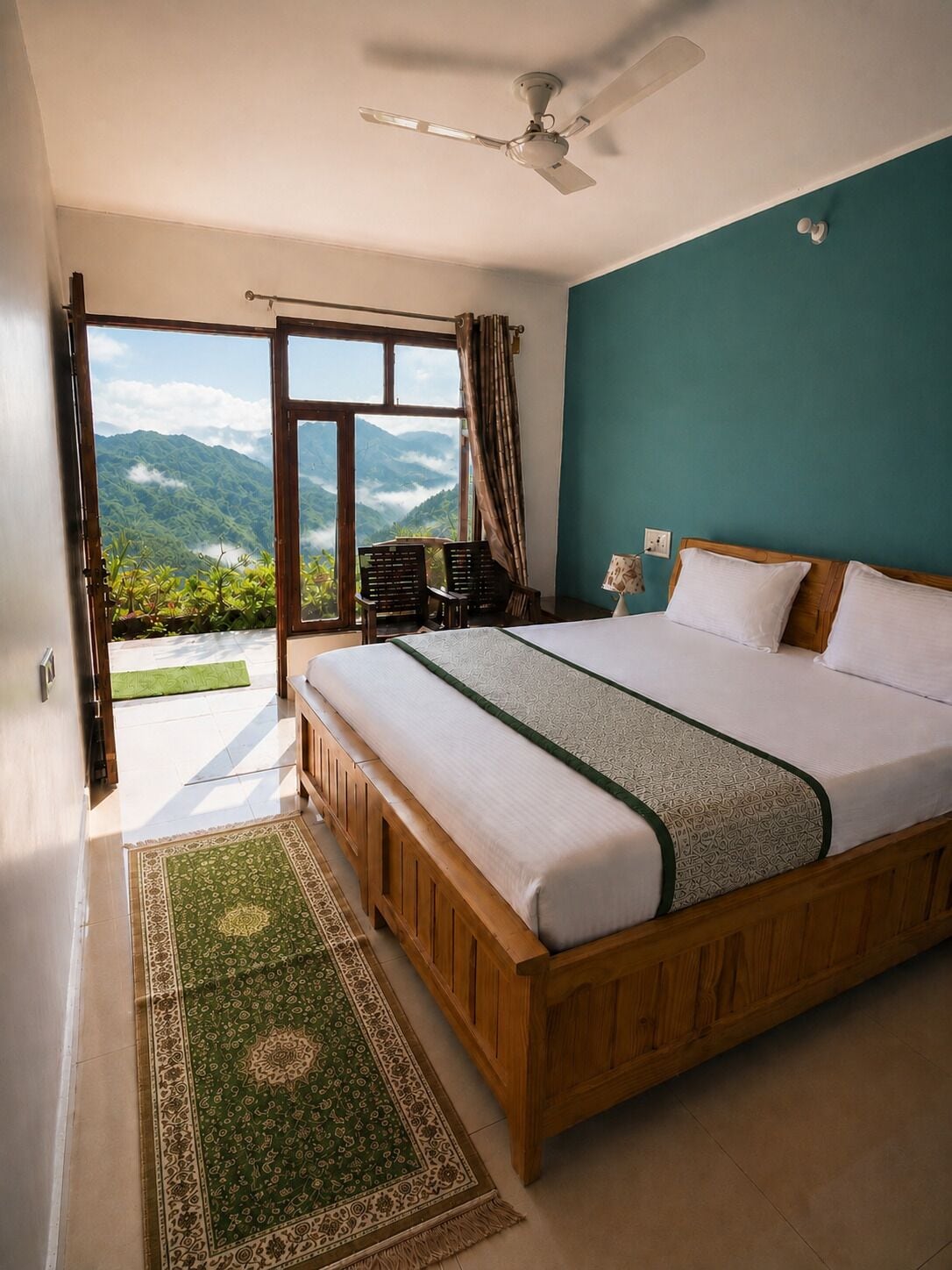 A bedroom with a large bed, a ceiling fan, a view of mountains through a window, and a green patterned rug.