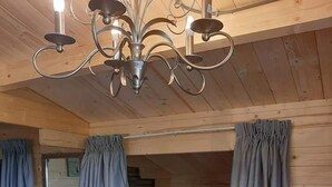 A wooden cabin room with a bed, curtains, a chandelier, a table with a potted orchid, and a small shelf with folded towels.