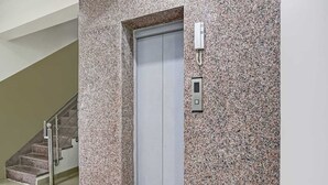 A modern elevator with a granite wall, a metal handrail, and a staircase with a metal railing. - The Vertigo (Noida)
