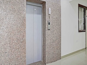 A hallway with a granite wall, a white elevator, and a wall-mounted intercom. - The Vertigo (Noida)