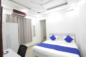 A hotel room with a bed, a desk, a chair, a wardrobe, a ceiling fan, and an air conditioning unit. - The Vertigo (Noida)