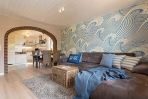 Ein Wohnzimmer mit braunem Sofa, blauen Kissen, einem gewebten Couchtisch und einem durch einen Bogen sichtbaren Küchenbereich.