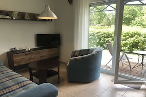 Ein Wohnzimmer mit einer Couch, einem Couchtisch, einem Fernseher auf einer Holzkommode und Blick auf Grün durch eine Glastür.