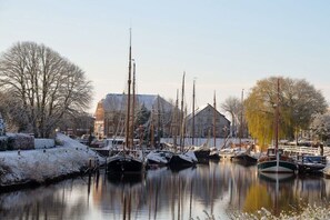 A canal with boats docked, a building in the background, and trees with snow on the ground. - Dream in Carolinensiel (Carolinensiel)