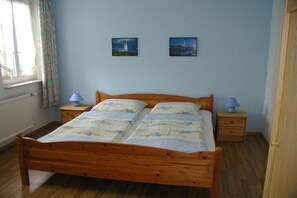 Ein Schlafzimmer mit einem hölzernen Bett, zwei Nachttischen, einem Fenster mit Vorhängen und zwei an der Wand befestigten Bildern.