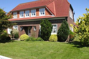 Ein zweigeschossiges Backsteinhaus mit rotem Ziegeldach, einer Veranda und einem gepflegten Rasen mit verschiedenen Sträuchern und Bäumen.