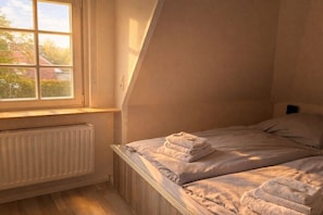 A bedroom with a sloped ceiling, a radiator, a window, and two beds with folded towels. - Langeneß in the house Achter de Dünen (Sankt Peter-Ording)