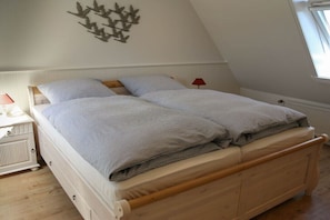 Ein Schlafzimmer mit schrägen Wänden, einem Bett mit Holzrahmen, zwei Kissen, einem Nachttisch mit Lampe und einer Wanddekoration.
