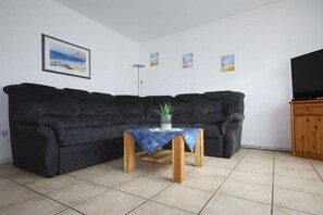 Ein Wohnzimmer mit einem schwarzen Sofa, einem Couchtisch aus Holz mit blauer Decke, einer Topfpflanze und einem Fernseher auf einer Holzkommode.
