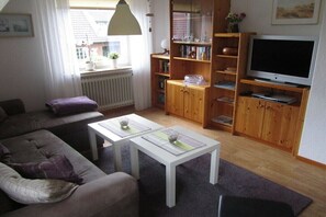 Een woonkamer met een bank, twee salontafels, een televisie en houten planken met boeken en decoratieve objecten.