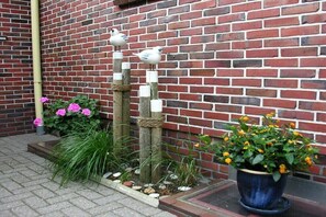 Eine Ziegelwand, eine hölzerne Vogelplastik und ein Blumentopf mit gelben Blüten.