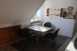 Een gezellig eetgedeelte met een tafel, stoelen, een radiator, een houten kast, een raam met gordijnen en een prikbord met papieren.
